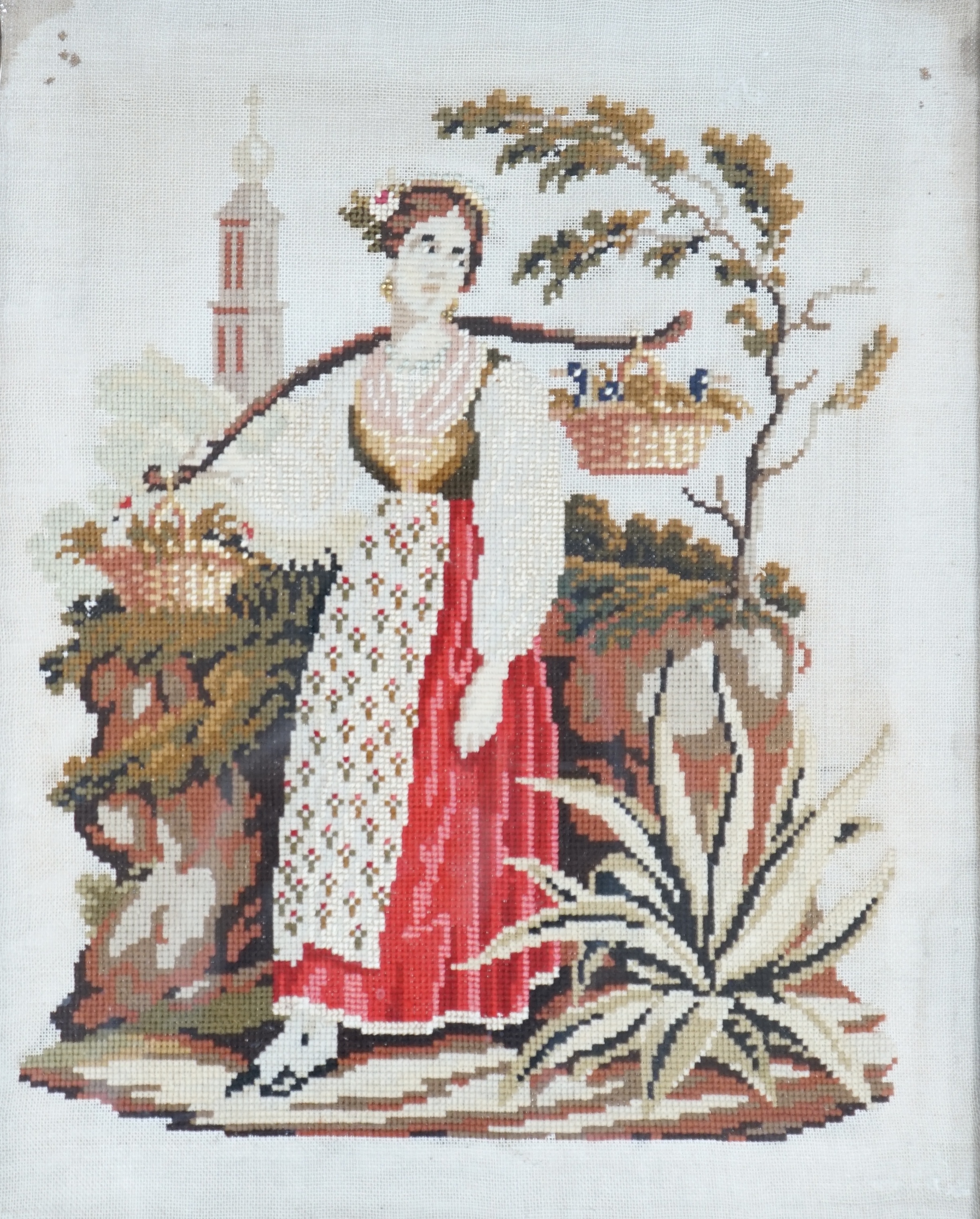 A framed 19th century Berlin wool worked embroidery of a peasant girl carrying panniers by a church, with the steeple in the background, 30cm x 38cm
